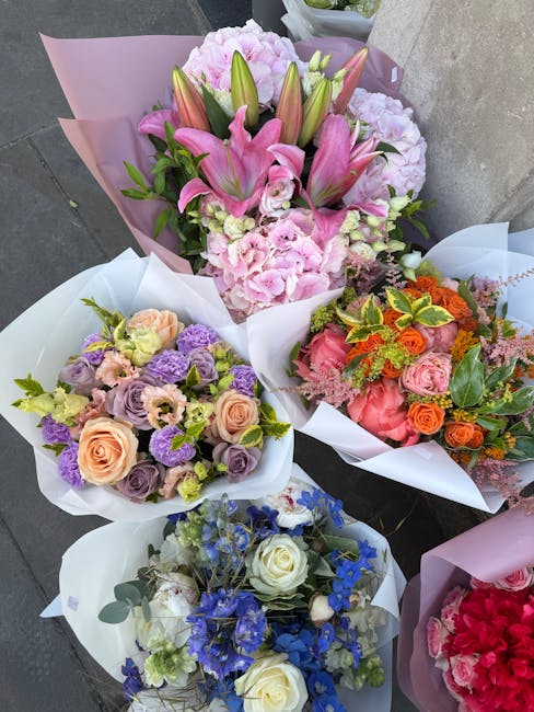 A collection of five beautifully arranged floral bouquets, each wrapped in glossy white and pastel pink paper, and displayed outdoors on a concrete surface. The central bouquet features a variety of p