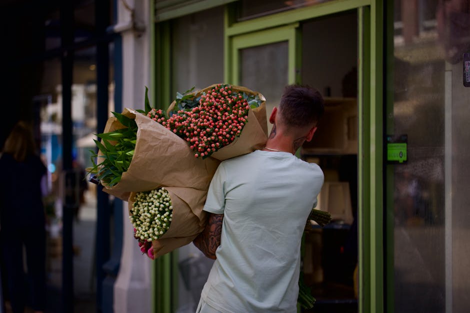 A person with short brown hair and a white t-shirt is holding a large bouquet of fresh flowers wrapped in light brown paper. The bouquet contains pink hypericum berries, white hypericum berries, green