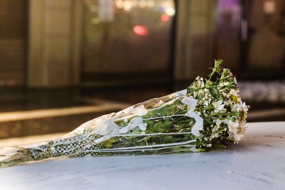 A bouquet of fresh flowers resting on a white surface outside a building with glass doors at night. The bouquet features white and light pink blossoms, including small roses and baby's breath, arrange