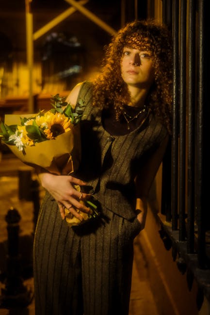 A young woman with curly brown hair and light skin holds a bouquet of fresh flowers wrapped in brown paper, featuring vibrant sunflowers, white daisies, and greenery. She is wearing a dark pinstripe v