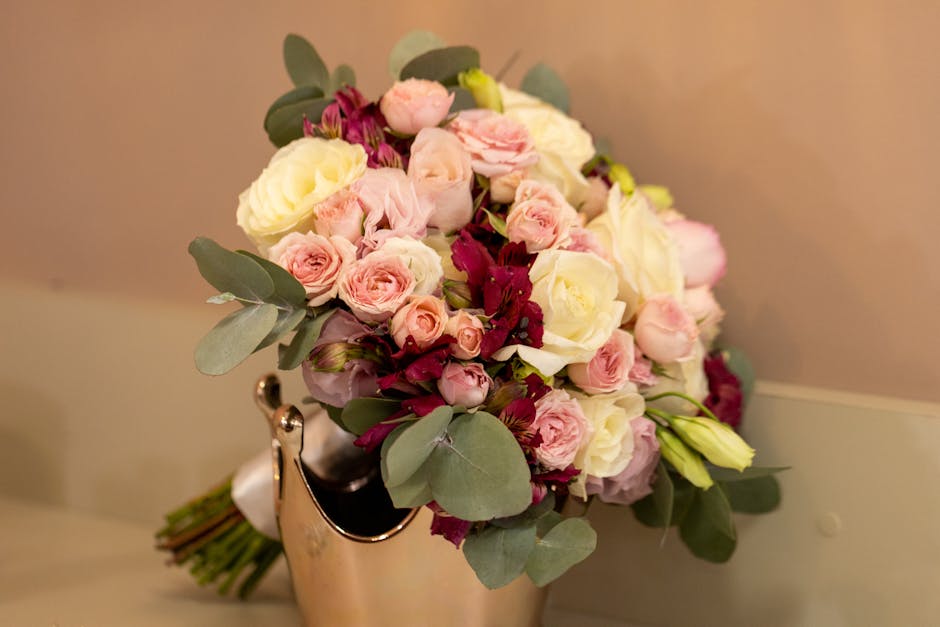 A bouquet of fresh flowers arranged in a rounded, compact style, featuring pale pink roses, white roses, and deep burgundy flowers, complemented by dusty miller eucalyptus leaves with rounded, silvery