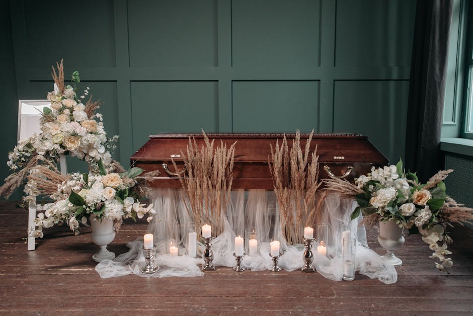 A floral arrangement for a funeral display featuring two large white ceramic urns filled with white roses, hydrangeas, and pale pink lisianthus, accented with greenery. Tall, feathery pampas grass sur