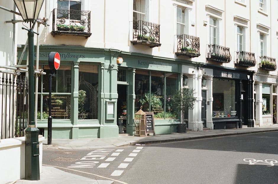 A view of a florist boutique located on a street corner in Notting Hill, London, with a pale green storefront displaying flower arrangements in the window. The shop's signage reads 'Kensington Flowers