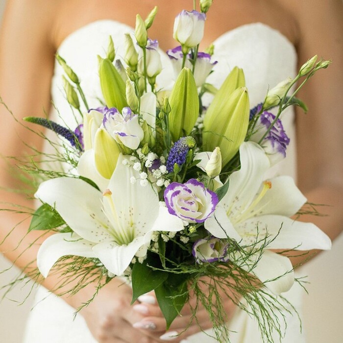 Seen through the lens of Notting Hill's vibrant spirit, this bridal bouquet is a symphony of white, green, and purple-expertly gathered by local florists familiar with the area's refined tastes. The composition is anchored by awe-inspiring Oriental lilies, their crisp white petals opening wide to reveal sun-dappled stamens and delicate spots-a floral focal point reminiscent of spring blooms in nearby Holland Park. Threaded alongside are ruffled lisianthus blooms, their edges gently tinged with lavender-purple, providing unexpected pops of colour and textural vitality. Royal purple spikes, likely Veronica sourced from London markets, rise between the more traditional blossoms, adding structure and a painterly effect. Wispy sprays of gypsophila dot the arrangement like flecks of morning mist, softening every line. Underpinning the flowers is a generous foundation of lush greenery-broad, glossy lily leaves and feathery asparagus fern-cascading over the bride's hands, which cradle the bouquet with understated grace, a trace of pale polish catching the light. The blurred white of the bride's strapless gown forms a glowing backdrop, framing the flowers in a haze of anticipation. Every detail-from buds to blooms, hues to arrangement-speaks to the careful artistry and local pride of Notting Hill floristry, crafted for couples seeking to mark their union with style and heartfelt tradition.