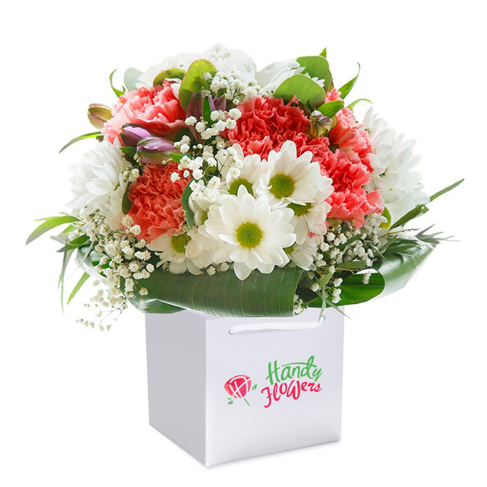 Standing tall in a sleek, white square gift bag, this floral arrangement captures the refined yet spirited style beloved in Notting Hill. The bouquet is a lush gathering of peach and coral carnations, identifiable by their abundant, frilled petals and plush texture, conveying both energy and grace. Interwoven among the carnations are pristine white daisies with animated lime-green centers, lighting up the arrangement with a cheerful contrast reminiscent of the neighborhood's vibrant terraces. Wispy sprigs of white baby's breath weave through the composition, imparting an ethereal finishing touch and soft, cloud-like lightness that mimics the ambiance of a calm day along Westbourne Grove. Nestled amongst these, hints of unopened buds, tinged with soft purple, quietly add mystery and depth, alluding to the hidden garden squares scattered throughout Notting Hill. Framing the flowers, a generous layer of deep, glossy green foliage forms a natural collar, their broad, shiny surfaces providing both visual structure and refreshing texture. The bag, specially designed for comfortable deliveries, features robust white ribbon handles for a polished, ready-to-gift look. This carefully composed scene, photographed in bright, even lighting, accentuates the vivid palette and textural variety of the blooms. Perfect for birthdays, thank-yous, or new beginnings, the bouquet brings together classic floral artistry and the personal touch of a local Notting Hill florist, ready to be delivered to West London's charming doorsteps.
