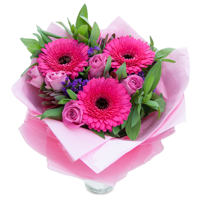 I see this composition as a blend of joyful colour and considered texture: three fuchsia-pink Gerbera daisies form the confident centre, each with numerous radiating petals and a dark central disk that anchors the arrangement visually, while a cluster of lavender-pink or dusty-rose roses nestle between them, their petals still tightly furled like a promise of bloom. Tiny sprays of purple statice - the kind you might find tucked into cottage gardens - thread through the bouquet, offering little pockets of cool colour and a feathery contrast to the fuller flowers. The greenery, generous and glossy, appears to be salal or eucalyptus, its leaves creating a deep, fresh backdrop that intensifies the pinks and carries a faint, uplifting green fragrance. The florist has wrapped the stems in a soft, matte pale pink tissue beneath a layer of sheer fuchsia-pink cellophane, slightly crinkled to catch light and add a subtle sparkle; these layers are artfully folded to present the blooms like a treasured gift. The visible rim of a clear glass vase at the bottom hints that the bouquet is ready to be arranged on a dining table or windowsill, where morning light - the kind that filters through Notting Hill terraces and touches Westbourne Grove shopfronts - will make the petals glow. This arrangement feels intimate and festive, suited to marking birthdays, anniversaries, or a thoughtful 'just because' gesture, bringing a sense of warmth and handcrafted care familiar to local recipients around Ladbroke Grove and Pembridge Villas.