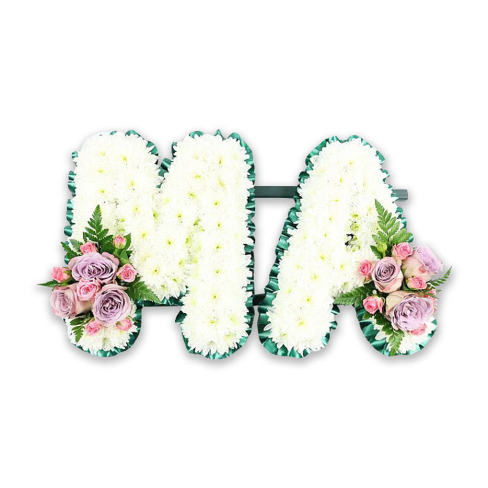 This image presents a refined funeral letter tribute that spells out MA in two strong, capital letters, carefully arranged so that every detail feels deliberate and respectful. The letters themselves are densely carpeted with pristine white chrysanthemums, each flower tightly packed until no gaps of the underlying frame can be seen. Their petals are short, soft, and slightly ruffled, giving the surface of the tribute a fluffy, cushion-like texture that invites the imagination to rest upon it. Light falls evenly across the arrangement, catching the whiteness of the blooms and creating delicate shading that helps define the shape of the M and the A. Encircling each letter is a deep green border, likely made from satin ribbon or finely placed foliage, neatly pleated to form a continuous, tailored edge. This rich green outline frames the snowy blooms with a clear line, in the same way the mature trees along Westbourne Grove frame the local streets with calm structure. At the base of the left side of the M and the right side of the A, two mirrored floral groupings add colour and emotional warmth. Here, soft lilac roses blend with gentle blush-pink roses and clusters of petite pink spray roses, their petals spiralling outward in layers that suggest tenderness and devotion. Light, feathery fronds of green fern extend slightly beyond the roses, introducing movement and a whisper of the natural world. The entire tribute rests against a plain white background, focusing attention on the craftsmanship and balance of colour. It feels like a bespoke design created to honour a beloved mother or Mama from the Notting Hill area, suitable for a funeral service, chapel of rest, or crematorium service, where family and friends gather to remember with love and dignity.
