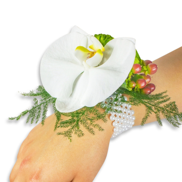 This elegant wrist corsage, photographed on a fair-skinned wrist against a clean white field, is centred on an immaculate white Phalaenopsis orchid whose wide, gently curving petals create a calm, architectural silhouette. The orchid's lip is a warm focal point - a bright yellow heart that graduates into a deeper orange at its core, giving the bloom a hint of sunlit warmth. Beneath and around the flower, airy asparagus fern provides a cushion of delicate, featherlike greenery that fans softly and catches the light, offering contrast to the orchid's smooth, porcelain texture. A neat cluster of hypericum berries sits to the upper right of the bloom; the berries move in tone from pale green to a blushing rose and finally to a rich cranberry, their glossy surfaces reflecting the surrounding light and adding a jewel-like accent. Near the berry cluster a small sprig of lime-green ruffled foliage adds a playful edge of texture. The whole composition is affixed to a classic multi-strand bracelet of small, shiny white pearls that wrap with gentle ease around the wrist, combining tradition with the freshness of living flowers. The photograph's soft, even lighting highlights the tactile differences - the cool, gliding pearl surface, the orchid's satiny petals, the fern's wispy fronds and the berries' taut sheen. It's a refined accessory ideal for weddings, proms, or an intimate celebration in Notting Hill or nearby Holland Park, where understated elegance and craftsmanship matter.