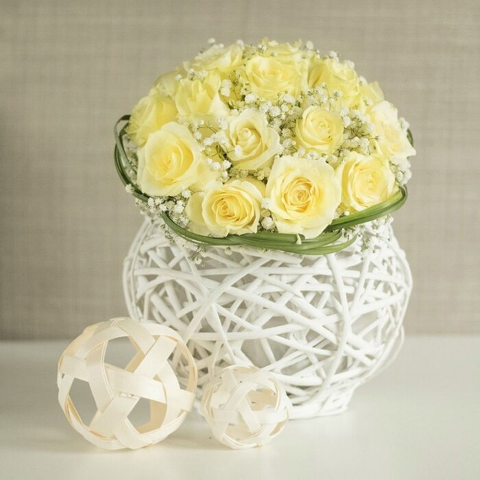 The image reveals a meticulously crafted wedding table centerpiece, the kind you might find gracing a long, candlelit table at a Notting Hill townhouse celebration. At its core sits a perfectly shaped dome of soft yellow roses, each bloom in full, plush openness, their petals layered in delicate spirals that transition from rich butter at the heart to paler cream at the tips. Interwoven among these roses are fine sprays of white baby's breath, thousands of tiny blossoms hovering like a delicate veil, softening the overall form and adding a gentle sense of movement, as though caught in a light breeze drifting up from nearby Portobello Road. A neat belt of smooth green foliage encircles the base of the flower dome, the leaves likely folded and wrapped with skilled precision to create a crisp edge that subtly separates the blooms from the container. The vase itself is a striking decorative piece: round, white, and fashioned from an intricate open weave that recalls modern wicker or rattan, its strands looping and crossing to form an airy, sculptural shell. Resting on the same spotless white surface, two pale woven spheres-one slightly larger than the other-sit in relaxed balance beside the vase, their open, geometric construction repeating the vase's texture and reinforcing the minimalist yet warm aesthetic. The background is a softly blurred neutral, perhaps a warm beige or light grey, creating a calm, gallery-like setting that places complete focus on the flowers and their tactile surroundings. The entire composition exudes quiet joy, thoughtful design, and a sense of fresh beginnings, perfectly suited to intimate weddings, bridal showers near Westbourne Grove, or tasteful home styling with flowers that feel both romantic and contemporary.