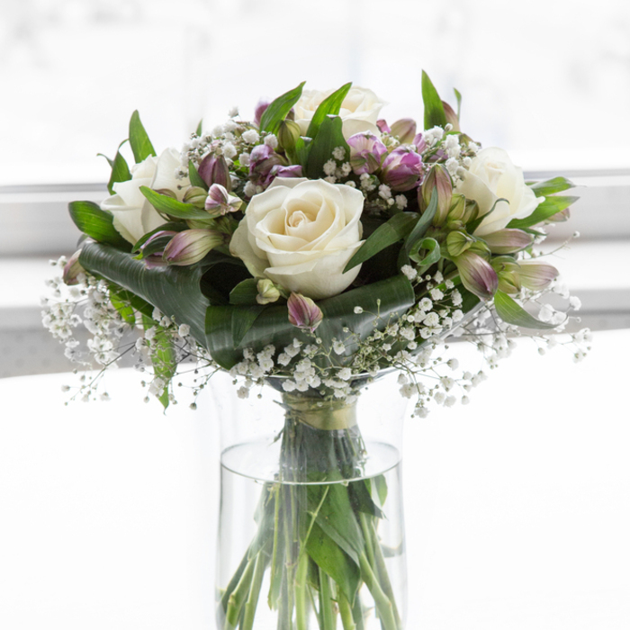 A refined bouquet of white and purple blooms is captured in a moment of stillness, arranged in a simple clear glass cylinder vase and illuminated by soft, even daylight, as though it rests on a polished table in a light-filled Notting Hill apartment. Several creamy white roses take centre stage, their petals unfolding in smooth, rounded layers that look almost luminous against the clean, bright backdrop. Woven between these focal blooms are elegant stems of alstroemeria, their petals shaded from soft lavender to deeper mauve, with hints of green at the base, adding gentle strokes of colour that never overwhelm the quiet harmony of the design. Airy sprigs of white baby's breath drift throughout the bouquet, filling spaces between the larger flowers and creating a fine, mist-like texture, reminiscent of early morning fog along nearby Holland Park. Lush green foliage forms a supportive frame, with glossy, broad leaves and slender green stems that provide structure and a sense of natural abundance. Through the transparent vase, straight green stems are visible in clear water, a simple detail that emphasizes the bouquet's freshness and authenticity. The background is a glowing, diffused white, with no visible objects, allowing the viewer to focus entirely on the interplay of soft whites, purples, and greens. The overall mood is one of calm, purity, and thoughtful remembrance, making this arrangement a fitting choice for expressing sympathy to families around Westbourne Grove or Bayswater, offering gentle comfort and a quiet, elegant tribute in times of loss.