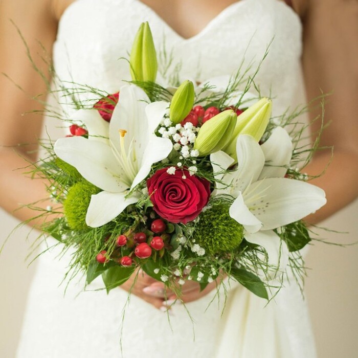 A luminous bridal bouquet comes to life in this close-up, cradled by the bride against the textured elegance of her strapless gown in the heart of Notting Hill. The focus falls on several sumptuous white oriental lilies, their petals arching open to golden, pollen-dusted stamens-a vision of purity and new beginnings. Intertwined are tightly furled, passionate red roses, their richly pigmented petals infusing the arrangement with romance and depth. Several vinous green lily buds, not yet unfurled, are artfully tucked between the larger blooms, promising a future in full blossom. Fine clusters of white baby's breath add an ethereal lightness, while glossy, cheerful hypericum berries lend distinct accents of red. Fuzzy, bright green trick dianthus spheres provide a unique tactile contrast, interspersed amid lush asparagus fern and deep green foliage, layering texture and fullness into the composition. The bouquet's fullness evokes the abundance of a local Notting Hill flower market; it seems as if you could almost catch the mingled scents of lilies and roses carried by a gentle Portobello breeze. With every stem thoughtfully chosen, this arrangement is at once classic and modern, echoing both tradition and the innovative artistry common to Notting Hill's florists. The bride's hands frame the bouquet with tenderness, capturing that fleeting, cherished moment as she prepares to walk down the aisle-her flowers as unforgettable as the streets outside.