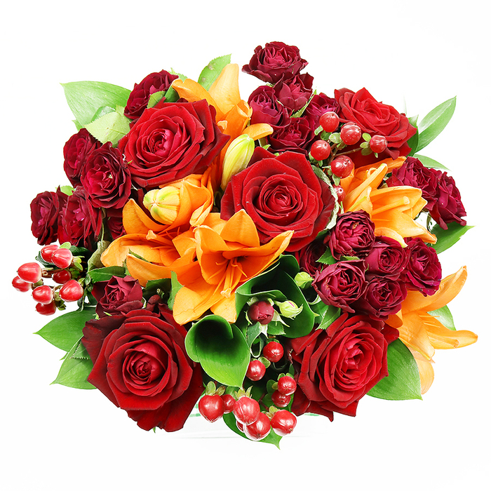 A sumptuous, tightly arranged bouquet photographed from above on a stark white background, this composition blends deep, romantic reds and fiery oranges with fresh green accents to striking effect. Large, fully bloomed deep red roses take centre stage - their thick, velvety petals layered into elaborate spirals that speak of classic romance and slow-moving passion. Scattered among them, bright orange Asiatic lilies introduce architectural trumpet shapes and the occasional speckled throat; stamens are clearly visible in some open flowers while other lily buds hold the promise of unfolding colour, offering a sense of temporal movement as if the bouquet might change over the course of an evening. Compact clusters of dark burgundy spray roses provide dense, textured pockets that intensify the jewel-toned core, while small, glossy hypericum berries - round and scarlet - add playful, reflective highlights and a contrasting firmness to the otherwise soft composition. Generous swathes of glossy green foliage, likely ruscus or salal leaves, push through between blooms, giving breath and structure and a cool counterpoint that prevents the warm palette from feeling too heavy. The soft, even studio lighting brings out the tactile differences - the satin sheen of lily petals, the matte opulence of rose velour, and the glossy surfaces of the berries - and the top-down viewpoint emphasises the bouquet's perfectly rounded form. Evocative of autumnal celebrations, romantic gestures or an elegant anniversary gift, this arrangement would sit beautifully as a centrepiece in a Notting Hill mews house or be passed as a heartfelt token along Ladbroke Grove toward Holland Park.