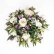 Captured from directly above against a pure white background, this bouquet has the calm, centred presence of a small garden gathered into a circle, the kind neighbours in Notting Hill might place on a hallway table during a time of mourning. At its heart, softly glowing white roses unfold in layers, their petals creamy and smooth, with subtle ivory and peach tones nestling deep in the centres. Some blooms are wide open, offering themselves fully to the light, while others remain in earlier stages, their cupped forms giving a sense of privacy and tenderness. Between them, slender stems of purple alstroemeria rise and curve, most of the blooms still in bud. Their pointed tips are washed in rich lavender and muted purple, gradually shifting into pale green and delicate pink near the stem, with a few buds just beginning to part enough to hint at the striped petals hidden inside. Scattered throughout, tiny blossoms of white baby's breath float like fine lace, contributing a gossamer softness that feels almost weightless, like the light drifting through a quiet side street off Portobello Road. Enveloping the arrangement, deep green foliage forms a lush collar, the leaves broad, glossy, and gently arched so that the bouquet appears round, full, and complete. The interplay of white, purple, and green creates a mood of tranquillity and sincerity, suitable for a condolence gift or a floral tribute at a memorial service. It carries the quiet message that someone is being held in thought and in heart, with the same gentle care a local Notting Hill florist would offer to families when they need comfort most.