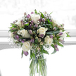 This image shows a calm and elegant sympathy bouquet standing in a clear, cylindrical glass vase, flooded with soft natural light that suggests a bright room in Notting Hill, perhaps looking out towards a quiet garden. At the core of the design, creamy white roses open slowly, each bloom full and rounded, with layers of smooth petals that create a feeling of tenderness and dignity. Intertwined among them are stems of alstroemeria in soothing shades of lilac, lavender, and pale green, their slender petals flaring gently outward, adding subtle movement and cool colour to the composition. Fine clusters of white baby's breath are scattered through the bouquet, forming tiny, cloud-like puffs that soften the transitions between roses and alstroemeria, and giving the whole arrangement a light, almost weightless quality. Deep green foliage, glossy and fresh, cradles the flowers from beneath, its varied shapes adding texture and a natural, garden-like fullness. The clear glass vase reveals the straight green stems and clean water inside, underscoring the sense of freshness and attentive floristry. Behind the bouquet, the background is a glowing, diffuse white, without any distracting detail, making the arrangement appear calm and uncluttered, almost like a moment of quiet reflection. The bouquet feels peaceful, respectful, and gently reassuring-the kind of thoughtful floral tribute a local might send to a home near Portobello Road or along Westbourne Park Road to express sympathy, remembrance, and unwavering support when someone is going through a time of grief.