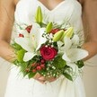 There's a sense of quiet anticipation in this fresh, hand-crafted bridal bouquet, captured in a luminous snapshot against a delicately patterned white wedding gown in Notting Hill. The visual center is formed by velvety white oriental lilies, their elegant petals unfurling to expose streaks of gold in their stamens, each blossom radiant with meaning. Woven between these, plush red roses nestle closely-every spiral a symbol of steadfast love and deep devotion. Playful clusters of shiny red hypericum berries and unique green button-like dianthus lend the design a touch of youthful joy and natural movement. Wisps of white gypsophila float, lending an airy, almost fairy-tale effect reminiscent of city gardens in early spring. Throughout, unopened green lily buds hint at new memories to come, while the entire bouquet rests on a verdant lattice of feathery ferns and broad glossy greens, embodying both abundance and calm. Soft daylight gently bathes the scene, highlighting not only the intricate craftsmanship of a Notting Hill florist but also the personal, heartfelt emotion held in the bride's gently clasped hands. This arrangement is more than florals-it's a story of love in bloom, ready to accompany her from iconic streets to a joyous celebration, and linger timelessly in memory.