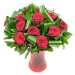 A lush, rounded bouquet of a dozen or more deep crimson roses fills the frame, each bloom at peak, velvety petals tightly spiralling toward a soft, wine-dark centre. The rich red tones are bold yet refined, catching a soft, even light that reveals delicate petal texture and subtle variations in hue. Surrounding the roses is a generous collar of glossy, dark green foliage that forms a dense, verdant backdrop. Narrow, flat leaves reminiscent of Ruscus or Salal create weight and depth, while broader, glossy Aspidistra-style leaves have been artfully folded and rolled into elegant curls and spirals. These sculpted green loops add a contemporary, architectural note, curving around the rose heads like protective frames. The stems sit in a tall, translucent ruby red glass vase with a faintly ribbed or twisted surface, vertical lines that catch and reflect light and echo the roses' passionate colour. Against a clean white background the composition reads as striking and intimate, the colours popping with gallery-like clarity. There is a quiet sense of fragrance suggested by the clustered blooms, a soft classic rose scent that feels both romantic and timeless. As a local Notting Hill arrangement, this would be equally at home on a dining table in a Holland Park flat or as a thoughtful anniversary gift delivered to a home off Portobello Road. The overall effect is elegant and composed, a modern take on a classic romantic bouquet that balances plush texture with precise, sculptural foliage and refined glassware.