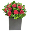 In crisp studio light a compact, contemporary bouquet reads like an exercise in elegant restraint: a tight cluster of rich, velvety red roses sits at the heart, each bloom opening in layered spirals of crimson that catch the eye with a satin glow. Those roses are cradled by broad, glossy green leaves that have been carefully curled and looped to create a rhythmic, sculptural backdrop; the foliage's waxy sheen and deliberate arcs give the composition a sense of movement while keeping the overall silhouette neat and geometric. The flowers are arranged low in a matte black cube, a boxy vase whose flat, modern surface grounds the arrangement and sets off the vivid reds and deep greens with a chic, urban contrast. Lighting is clean and steady, catching highlights on petal edges and the glossy leaf curves so the roses glow almost as if lit from within, and the whole piece conveys a tactile contrast between soft, silken petals and the firmer, almost tropical texture of the greenery. There's a subtle rose scent that feels neatly contained-classic and reassuring-tempered by the green freshness of the leaves. As a floral study this bouquet suggests intimate celebration: an anniversary dinner in a Notting Hill mews, a romantic note tucked into a Portobello Road gift, or a minimal but heartfelt table accent for a Westbourne Grove apartment. Described to someone who cannot see it, the bouquet reads as both dramatic and composed, a small statement of affection and thoughtful design rooted in local urban living.